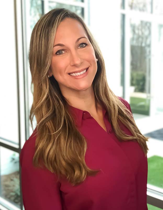 Professional headshot of a woman with long light brown hair wearing a burgundy blouse, smiling in front of large windows with natural light.